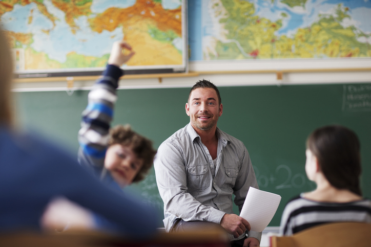 Teacher in classroom looking at students with their hands up.