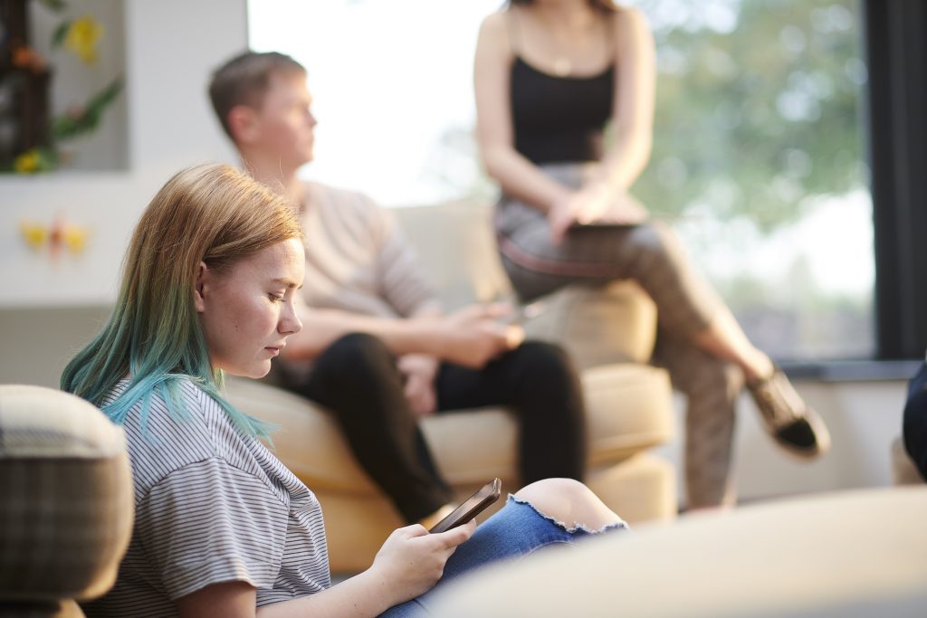 Young woman with long hair sits on floor with back against couch looking at her phone. Blurred out behind her are a yound man and woman sitting on a couch talking.