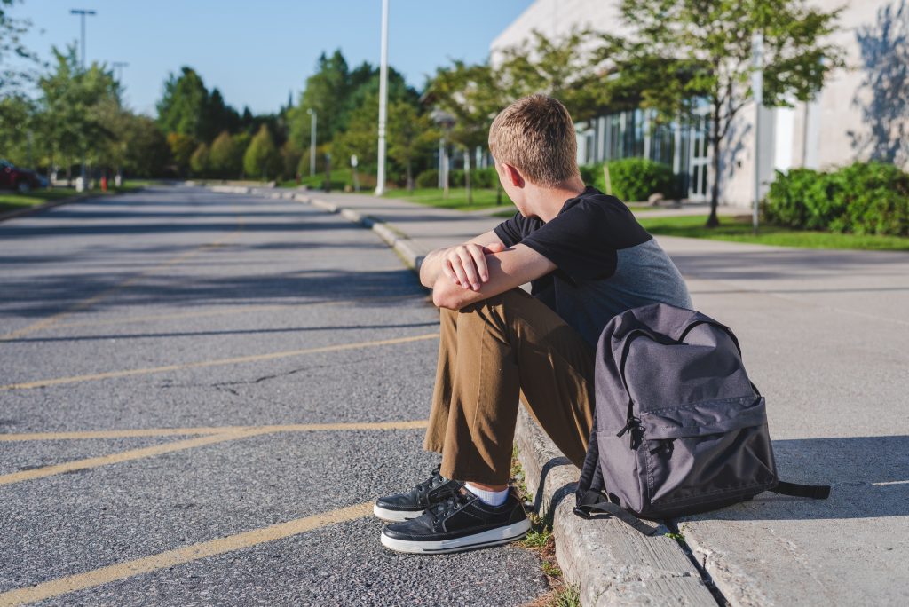 A student sits on the curb of a road with arms crossed and resting on his knees, looking away.