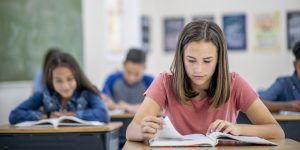 A high school student sits at their desk, with their textbooks open in front of them as they read. Other students can be seen behind them, doing the same. They are all dressed casually and are focused on the task.