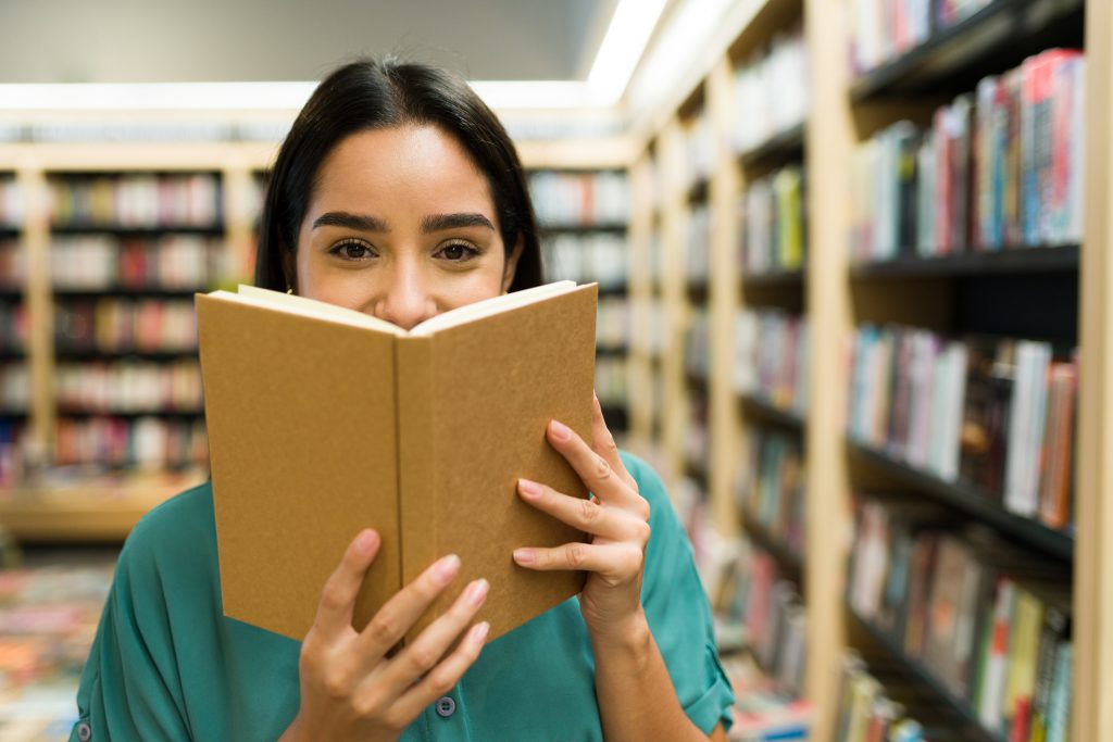 Happy young woman smiling while hiding behind a book held open up to her face, with bookshelves surrounding her.