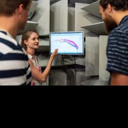 A person with long blonde hair and a colourful dress explains a line graph displayed on a monitor to two other people.