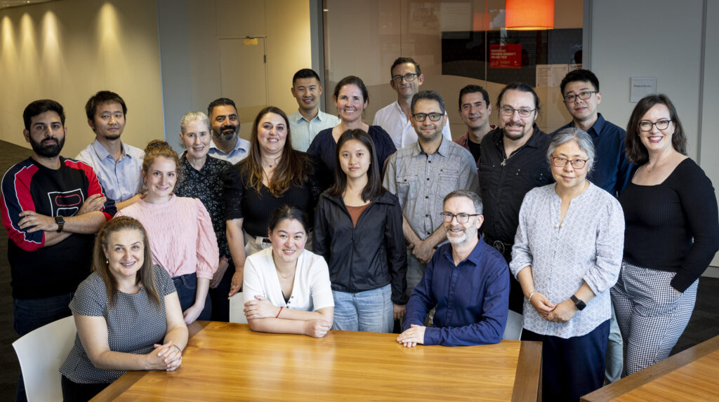 A large team of scientists gathered around a wooden table, some sitting, some standing, all smiling at the camera.