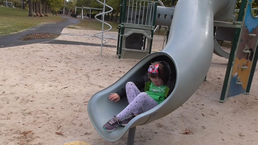 Girl’s body emerges at the end of a closed slide. Her body is curled over to fit through slide. She is using her hands and legs to wriggle to the end.