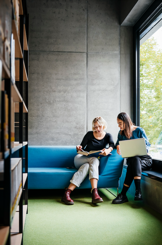 Researchers sitting in a library. One has a book, one is using a laptop. Sitting on a blue couch by a window on green carpet.