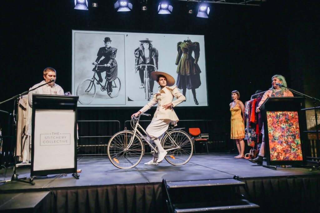A person wearing a historical bike riding outfit sits on a bicycle on a stage in front of a screen showing examples of similar clothing.